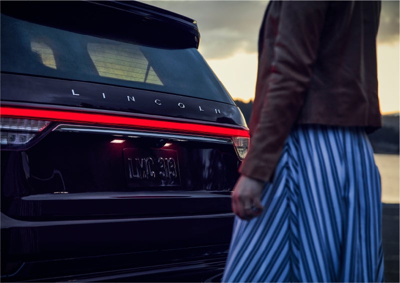 A person is shown near the rear of a 2023 Lincoln Aviator® SUV as the Lincoln Embrace illuminates the rear lights | Al Packer Lincoln in West Palm Beach FL