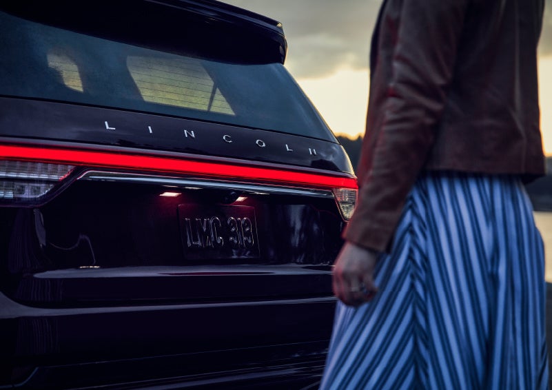 A person is shown near the rear of a 2024 Lincoln Aviator® SUV as the Lincoln Embrace illuminates the rear lights | Al Packer Lincoln in West Palm Beach FL