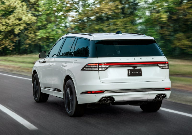 A white 2026 Lincoln Aviator® SUV is being driven away down a road lined with colorful trees. | Al Packer Lincoln in West Palm Beach FL